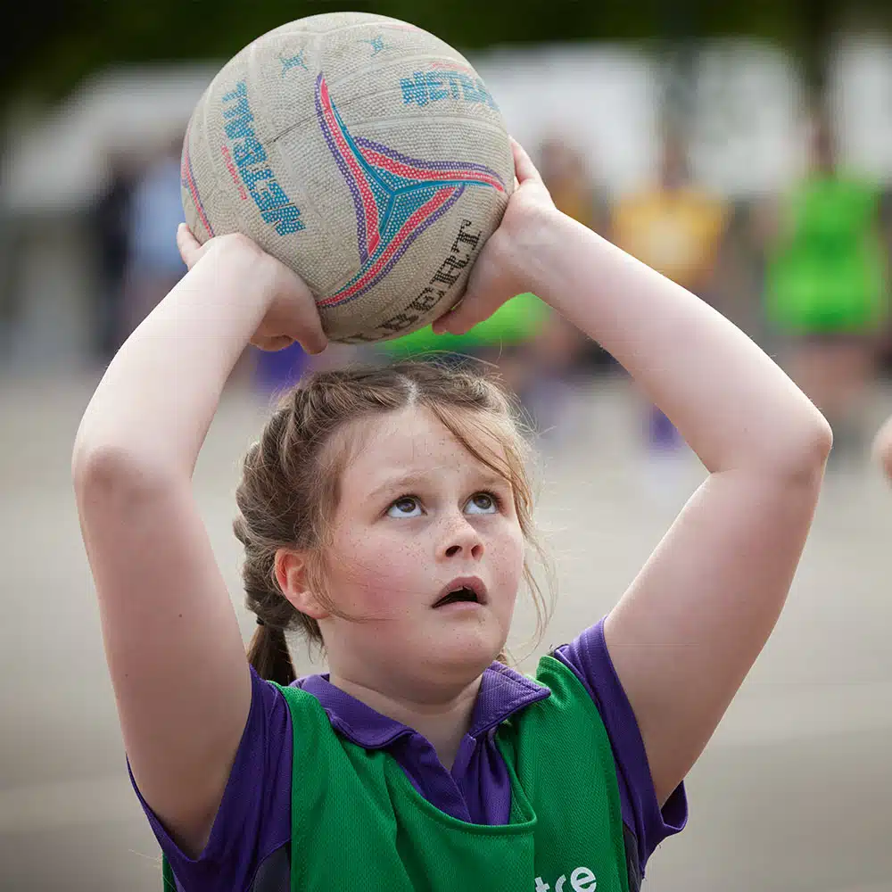 Young girl in a green and purple sports uniform, preparing to shoot a netball outdoors during her PGL Adventure Holiday. She is looking upwards, holding the ball above her head with both hands.