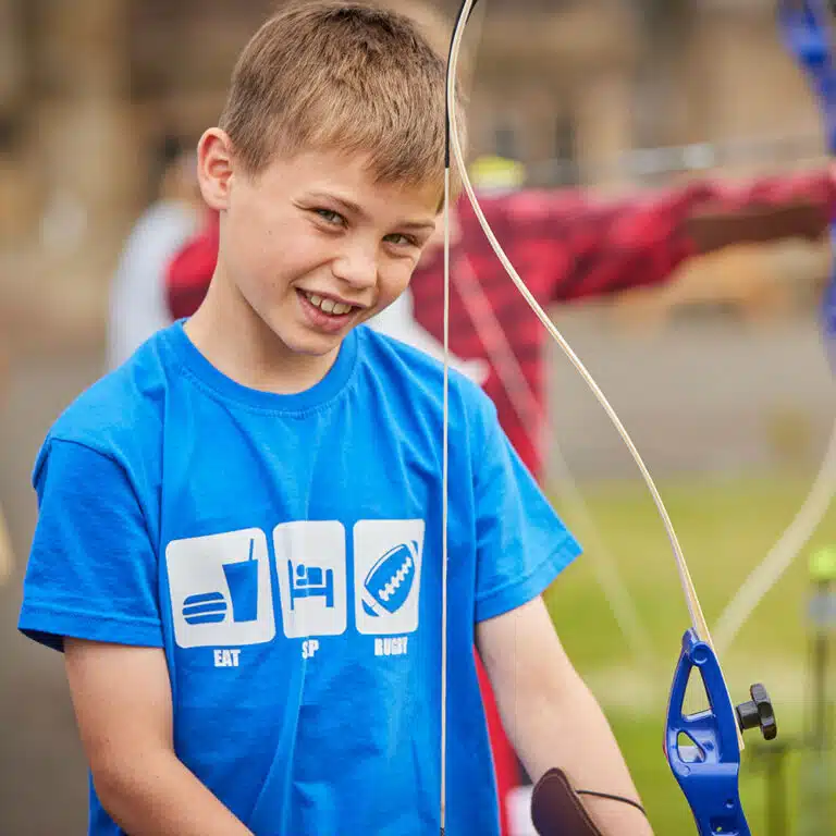 A young boy wearing a blue shirt holds a bow, standing outdoors and smiling during his PGL Adventure Holidays experience.