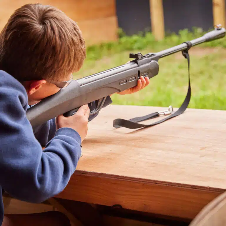 A person with short hair wearing goggles aims a rifle at a shooting range, resting the weapon on a wooden surface during an exciting activity at PGL Adventure Holidays.