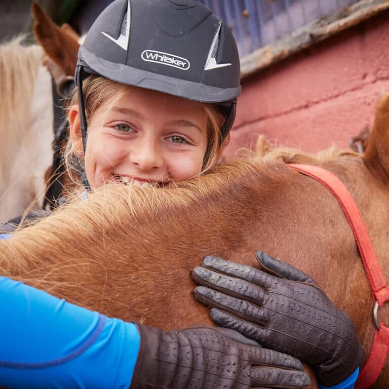 A young person wearing a riding helmet and gloves is hugging a brown horse in an outdoor stable area, reminiscent of the heartwarming moments often experienced on PGL Adventure Holidays.