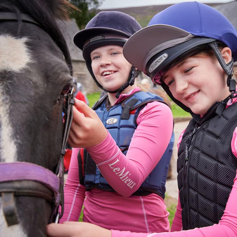 Two young people wearing helmets and equestrian vests pet a horse. They are smiling and appear to be outdoors near a stable, enjoying their time on a PGL Adventure Holiday.