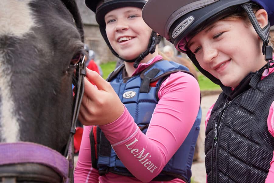 Two young people wearing helmets and equestrian vests pet a horse. They are smiling and appear to be outdoors near a stable, enjoying their time on a PGL Adventure Holiday.