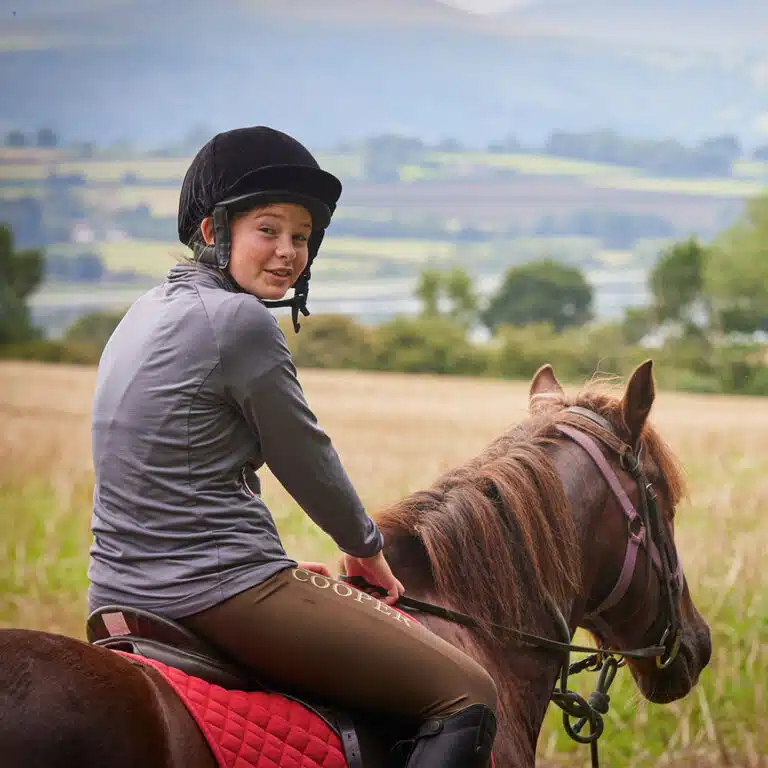 A person wearing a riding helmet and gray jacket is sitting on a brown horse with a red saddle pad. They are outdoors in a field with hills and trees in the background, enjoying the thrills of PGL Adventure Holidays.