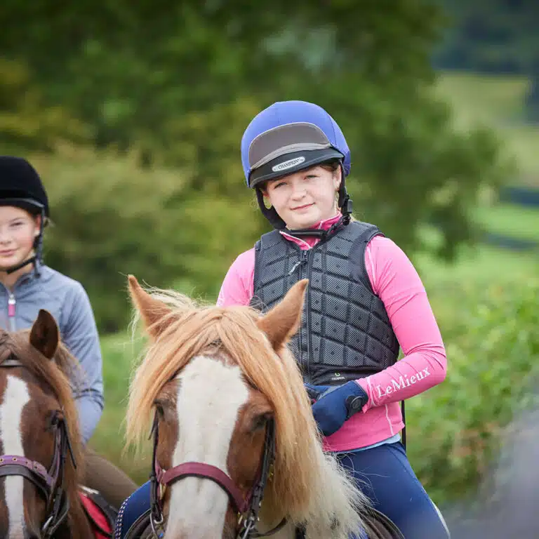 A young person wearing a helmet and protective vest rides a brown and white horse outdoors, with another rider visible in the background, enjoying the experience as part of their PGL Adventure Holidays.