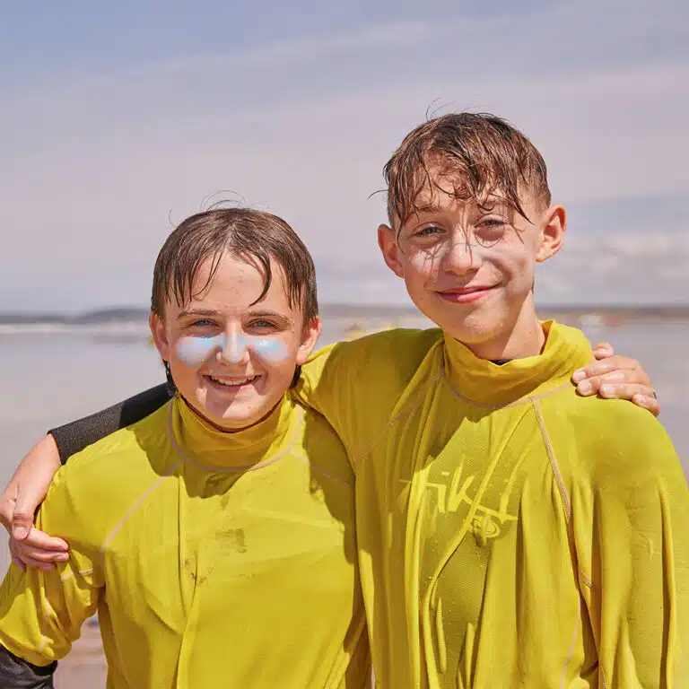 Two children with wet hair and yellow high-neck swim shirts stand arm-in-arm on a beach, smiling at the camera during their PGL Adventure Holidays. Both have white sunscreen smeared on their faces.