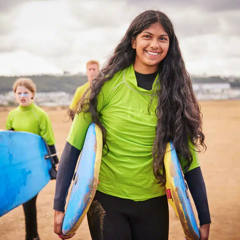 A person with long hair in a green shirt carries surfboards, embodying the spirit of PGL Adventure Holidays. Two other people in green shirts and wetsuits are in the background, ready for their next thrill.