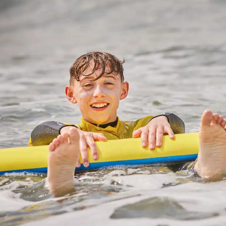 A child in a wetsuit floats in water holding a yellow pool noodle, smiling at the camera during their exciting PGL Adventure Holidays.
