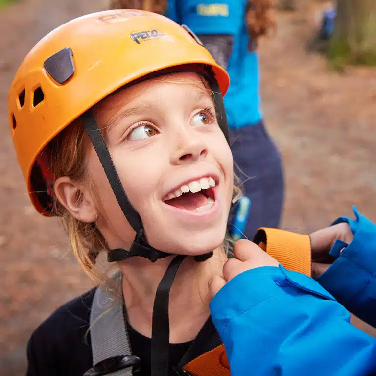 A child wearing an orange helmet smiles while another person adjusts their harness. They are outdoors, possibly preparing for an adventure with PGL Adventure Holidays.
