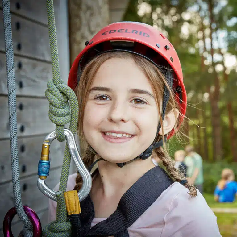 A smiling child wearing a red helmet and climbing harness stands next to ropes and carabiners, ready for an exciting day with PGL Adventure Holidays. Trees and a building are visible in the background, adding to the outdoor adventure vibe.