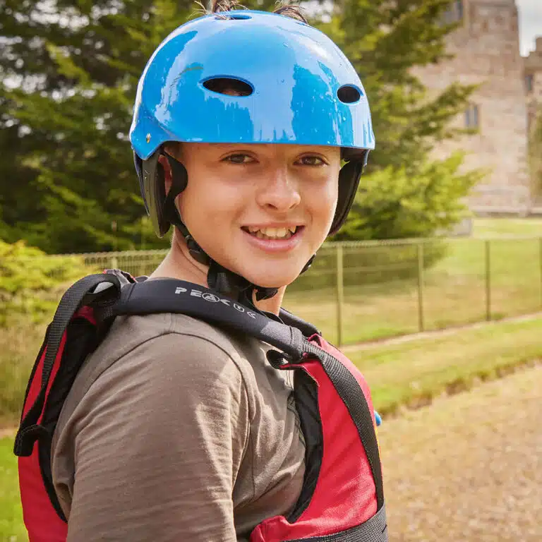 A person wearing a blue helmet and a red life jacket smiles outdoors with trees and a building in the background, enjoying their PGL Adventure Holidays experience.