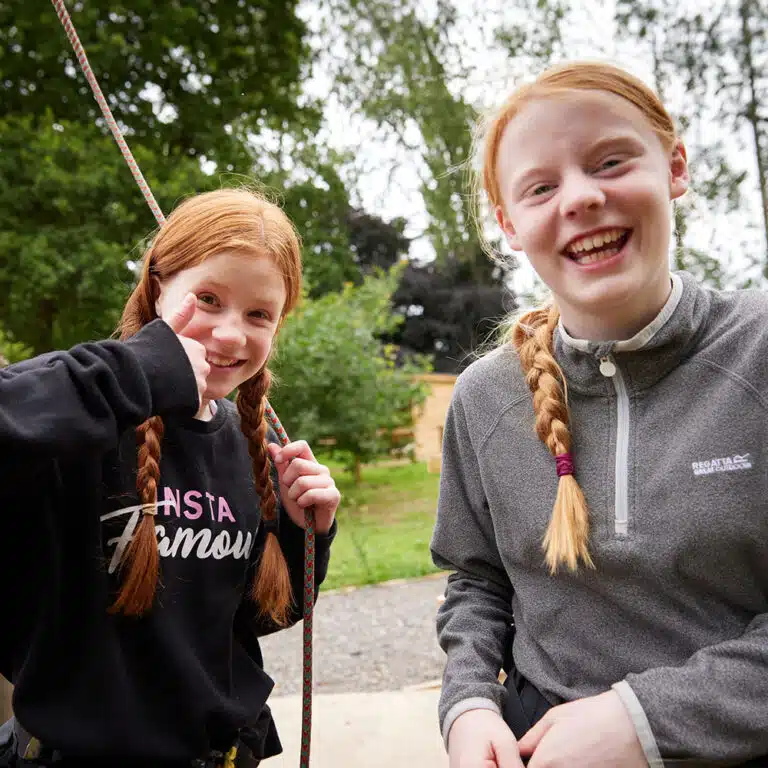 Two girls with red hair and braids smile outdoors, one giving a thumbs-up gesture. Both are wearing casual clothing and are surrounded by greenery and trees, enjoying their time on a PGL Adventure Holiday.