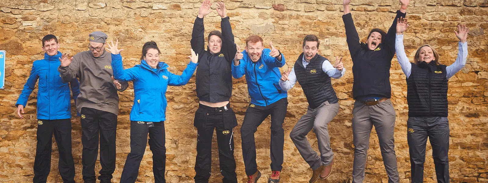 A group of eight people wearing various outdoor gear enthusiastically jump in front of a stone wall, embodying the spirit of PGL Adventure Holidays. Four are wearing blue jackets, and four are in darker jackets.
