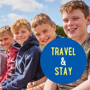 Four boys sit close together outside, smiling at the camera, with a blue circle in the foreground containing the text "TRAVEL & STAY.