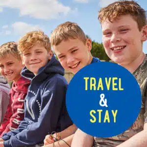 Four boys sit close together outside, smiling at the camera, with a blue circle in the foreground containing the text "TRAVEL & STAY.