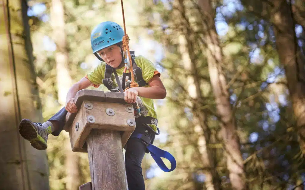 A young boy climbing a wooden structure at a high ropes course, wearing a blue helmet and harness, concentrating on his next step.