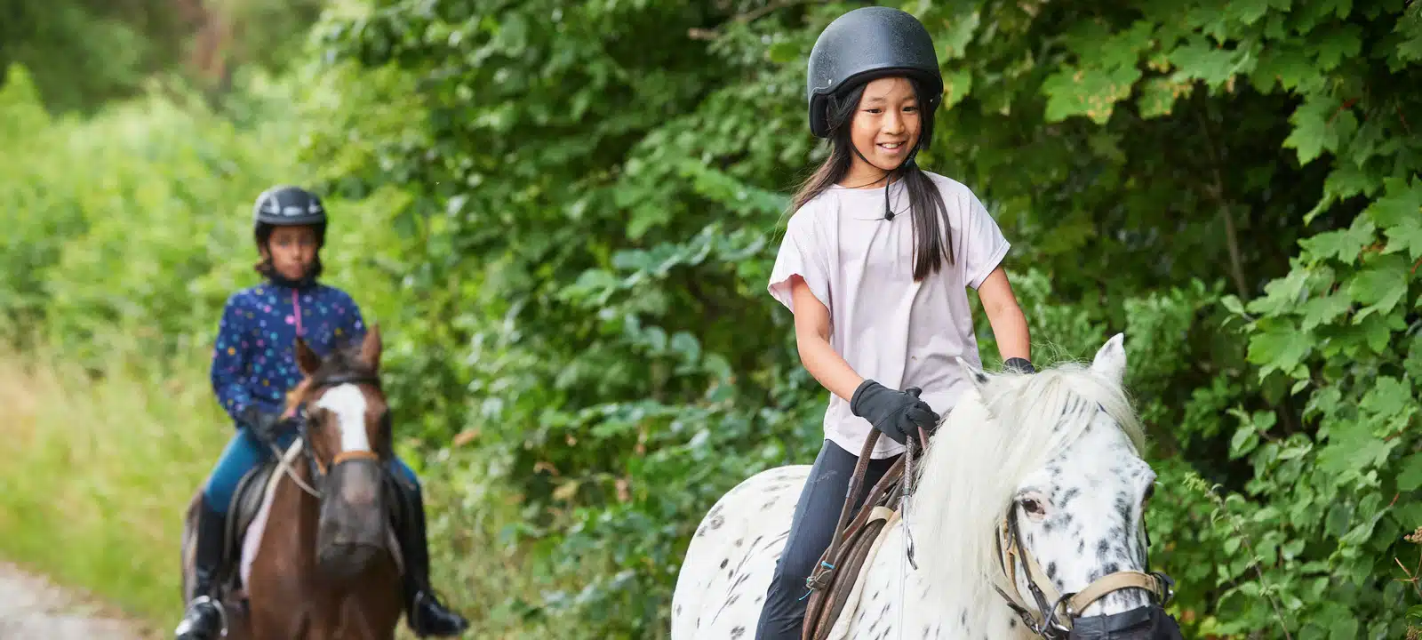 Two children enjoying a PGL Adventure Holidays ride on horses along a forest trail, with the front child smiling and leading on a white spotted horse.