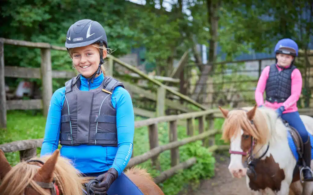 Young girl in blue riding gear smiling on a horse at PGL Adventure Holidays, with another child on a horse in the background.