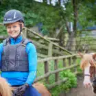 Young girl in blue riding gear smiling on a horse at PGL Adventure Holidays, with another child on a horse in the background.