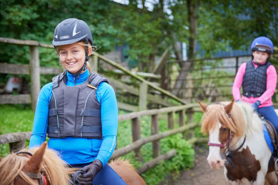 Young girl in blue riding gear smiling on a horse at PGL Adventure Holidays, with another child on a horse in the background.
