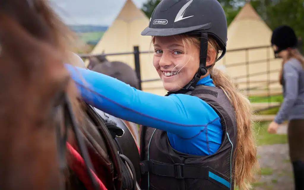Young girl in a helmet and blue shirt smiling while adjusting a horse's saddle at an outdoor riding camp.