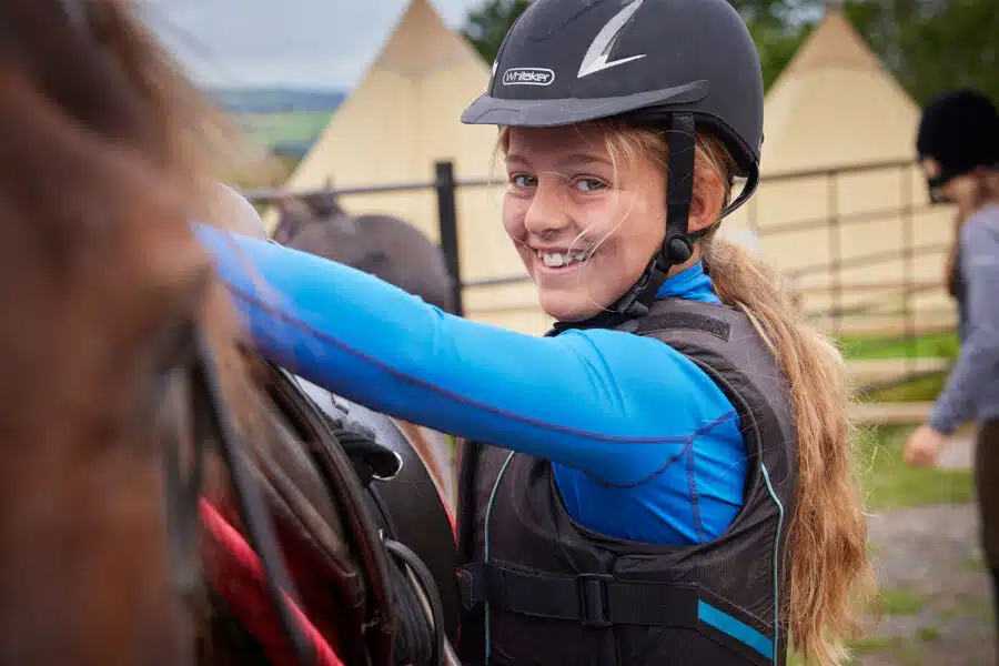 Young girl in a helmet and blue shirt smiling while adjusting a horse's saddle at an outdoor riding camp.