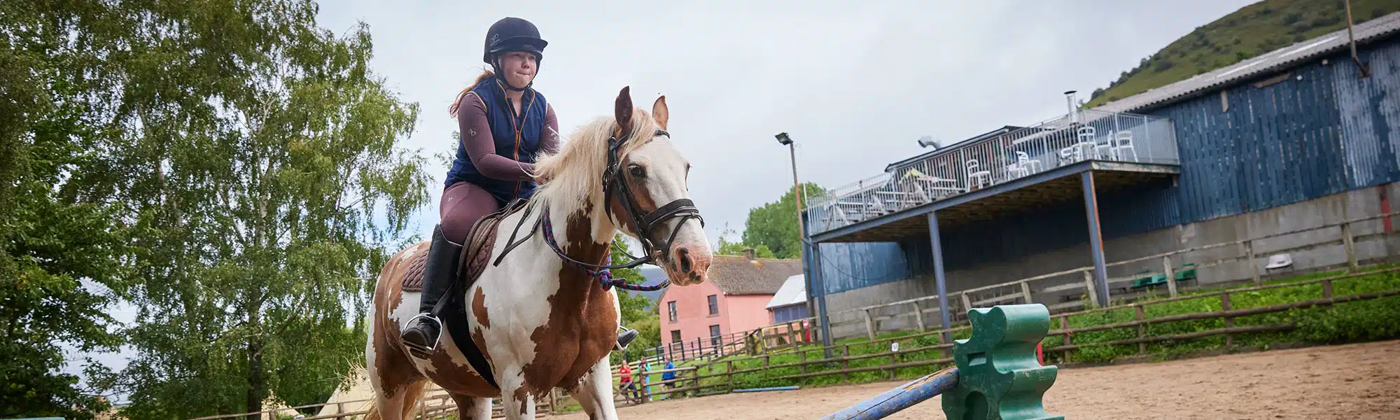 A woman in a helmet riding a white horse in an outdoor arena near colorful buildings and green hills.