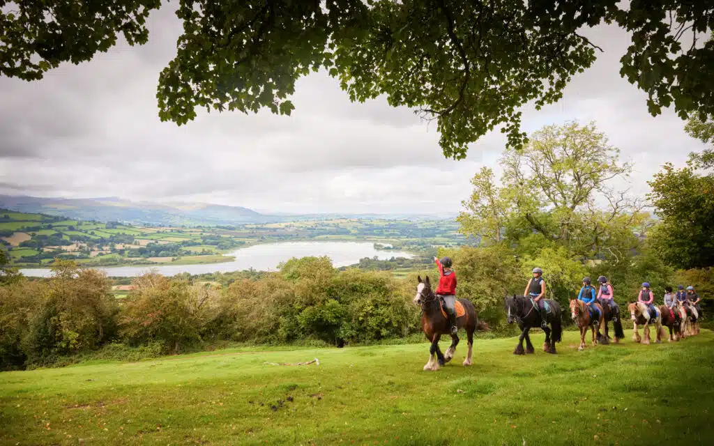A group of riders on horses trekking through a green hillside with a distant lake and overcast sky in the background during their PGL Adventure Holidays experience.