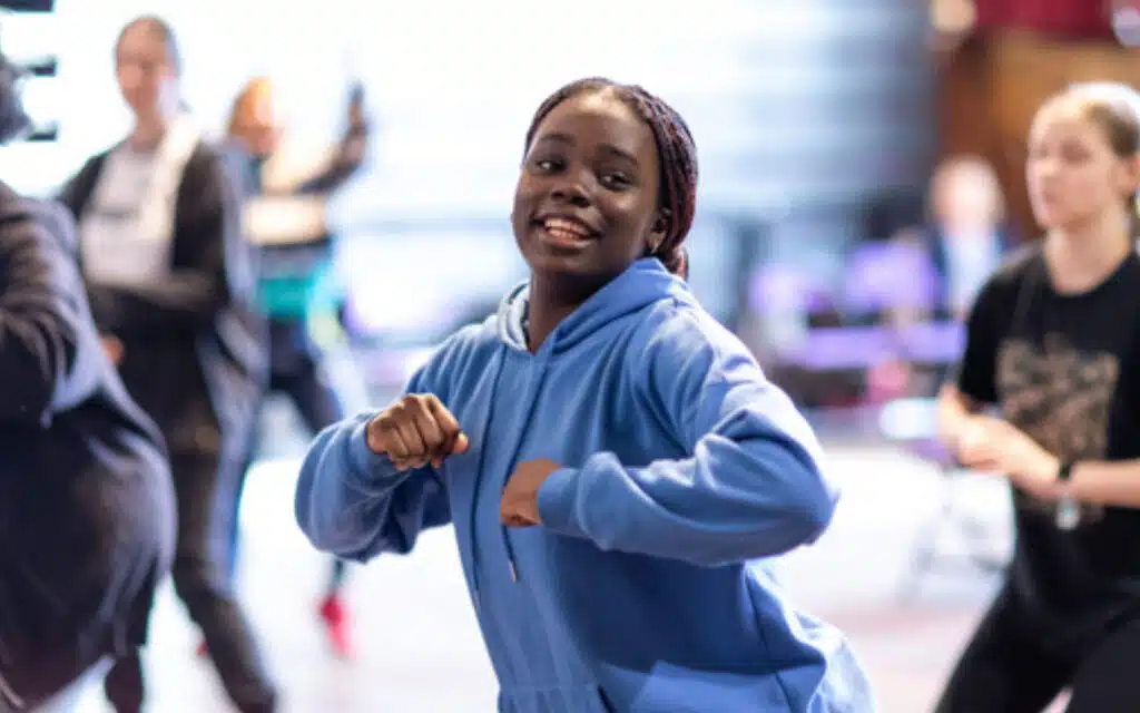 A young woman in a blue hoodie smiles while dancing in a dance studio during PGL Adventure Holidays, with others in the background.