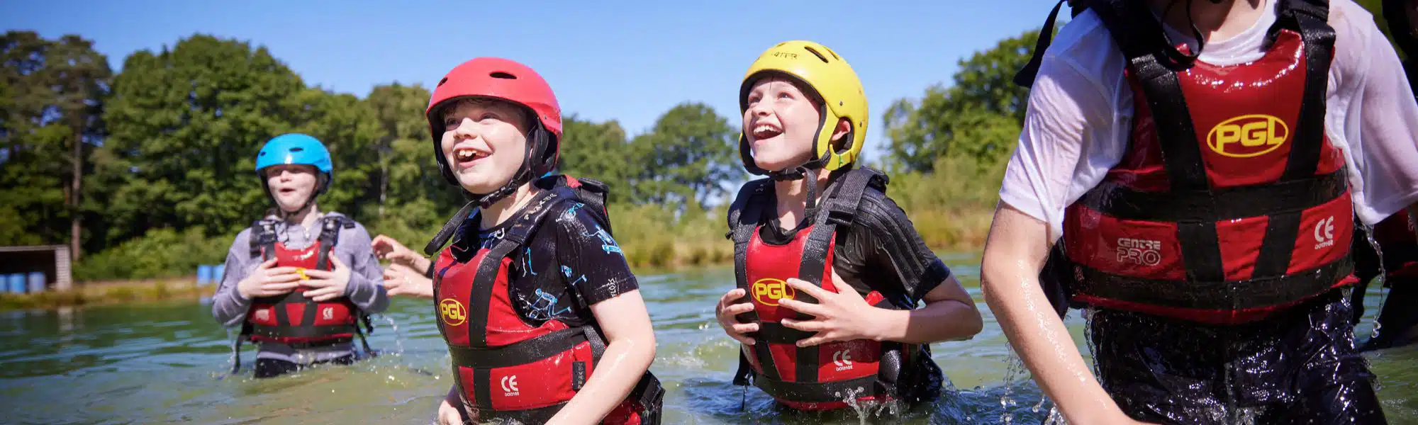 Four children wearing helmets and life vests are enjoying themselves in a body of water on a sunny day during their PGL Adventure Holidays.