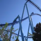 A close-up view of blue steel roller coaster tracks with loops and turns against a clear blue sky, surrounded by some greenery.