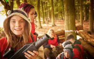 A group of young people in a forest are seen engaging in a laser tag activity, as part of their PGL Adventure Holidays. In the foreground, one girl holds a laser tag gun and smiles at the camera.