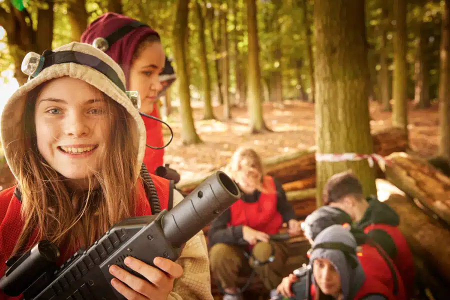 A group of young people in a forest are seen engaging in a laser tag activity, as part of their PGL Adventure Holidays. In the foreground, one girl holds a laser tag gun and smiles at the camera.