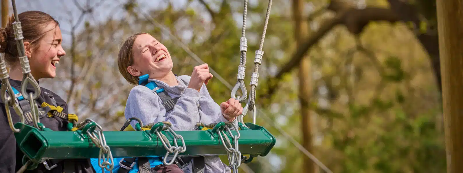 Two young people wearing harnesses laugh and enjoy themselves while seated on a suspended green swing with trees in the background, making memories thanks to PGL Adventure Holidays.