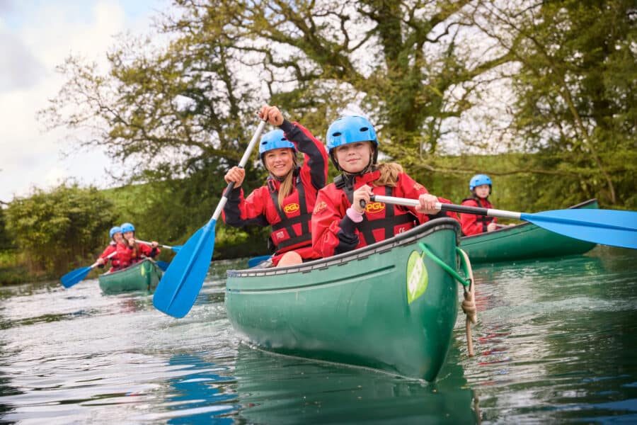A group of people wearing helmets and life jackets paddle green canoes on a calm river surrounded by trees, enjoying a PGL Adventure Holiday.