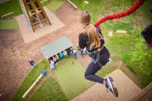 A person with long hair, wearing a harness, jumps off a platform during a PGL Adventure Holidays quick jump activity while a group of people watch from below.