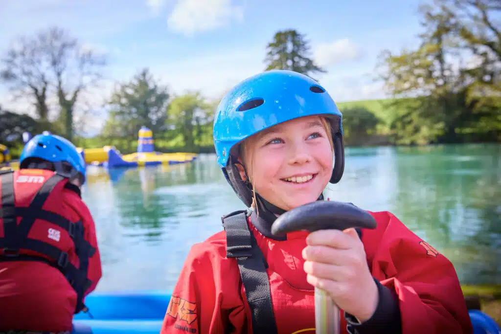 Two children in red jackets and blue helmets are participating in a water activity by a lake during their PGL Adventure Holidays. The child in the foreground is smiling and holding a paddle, while an inflatable structure is visible on the water.
