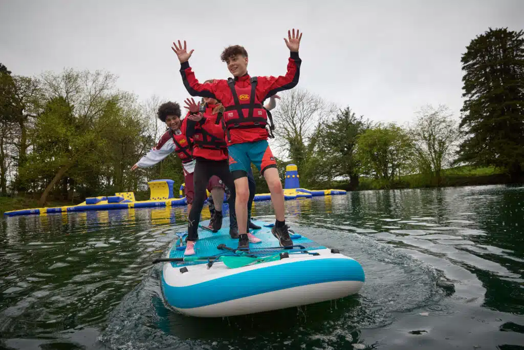 Three people in life jackets balance on a large paddleboard on a lake during their PGL Adventure Holidays. Trees and water inflatables are visible in the background.