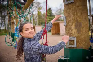 A person partakes in a PGL Adventure Holidays experience by climbing a wooden pole with safety ropes at an outdoor adventure park. Wearing a harness and gripping a metal handle on the pole, they look confidently at the camera.