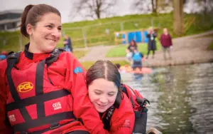 Mother and daughter wearing red life jackets sit near a body of water.