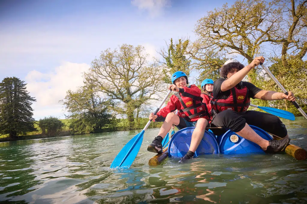 Three people in helmets and life jackets paddle a makeshift raft made of barrels and logs on a calm body of water, part of their exhilarating PGL Adventure Holidays experience, with trees providing a scenic backdrop.