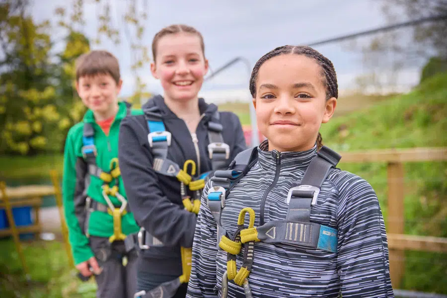 Three children wearing safety harnesses stand in line outdoors, smiling at the camera at PGL Adventure Holidays, with greenery and a fence in the background.