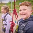 Two children in safety harnesses are at an outdoor activity area. The boy in the foreground smiles while holding a red rope with carabiners. The girl in the background is slightly out of focus.