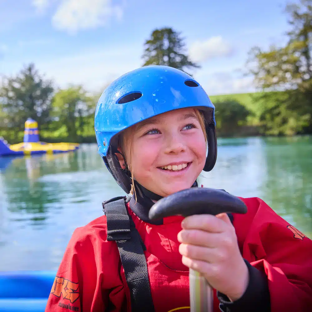 A child wearing a blue helmet and red jacket smiles while holding a paddle on a lake with trees and an inflatable structure in the background.
