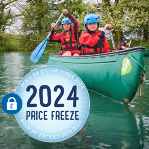 Two children in helmets and life jackets paddle a green canoe on a calm lake. A circular overlay promotes a "2024 Price Freeze" offer if booked by October 31st, 2024.