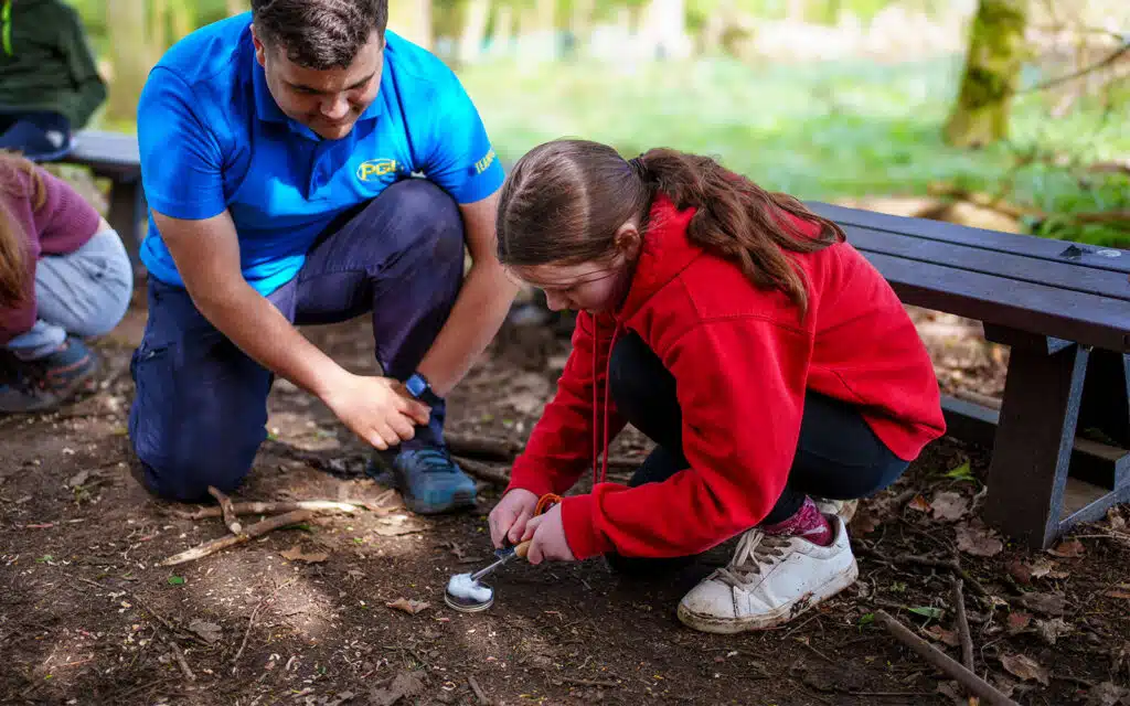 A man in a blue shirt, reminiscent of PGL Adventure Holidays, helps a child in a red sweatshirt using a spoon to dig in soil beside a wooden bench outdoors.