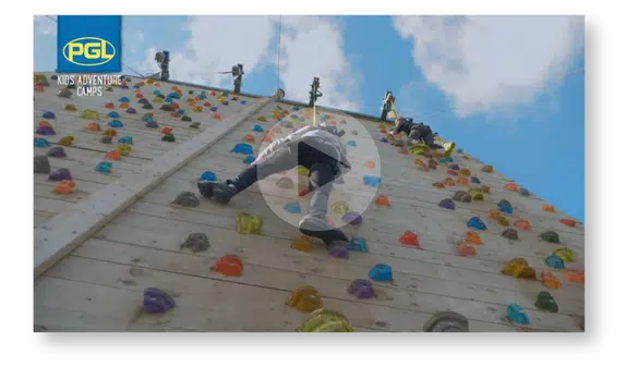 Children enjoy the thrill of climbing a large outdoor wall with colorful handholds under a blue sky, experiencing the excitement of PGL Adventure Holidays.