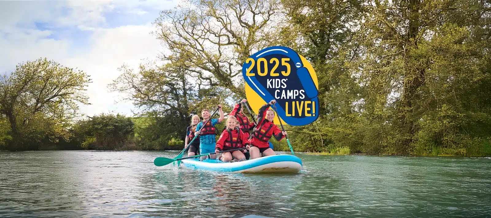 A group of children wearing life jackets paddle a large inflatable board on a calm river, with a "2025 Kids' Camps Live!" sign above them.