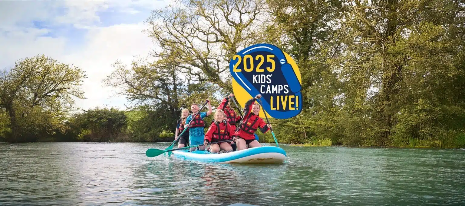 Children in red life vests paddle on a large board in a river, surrounded by trees. A circular "2025 Kids' Camps Live!" logo is in the upper center of the image.