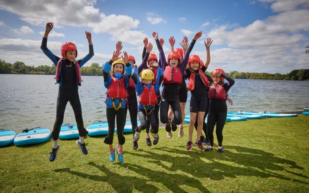 A group of people wearing helmets and life jackets jump excitedly by a lake with paddleboards on the grass behind them.