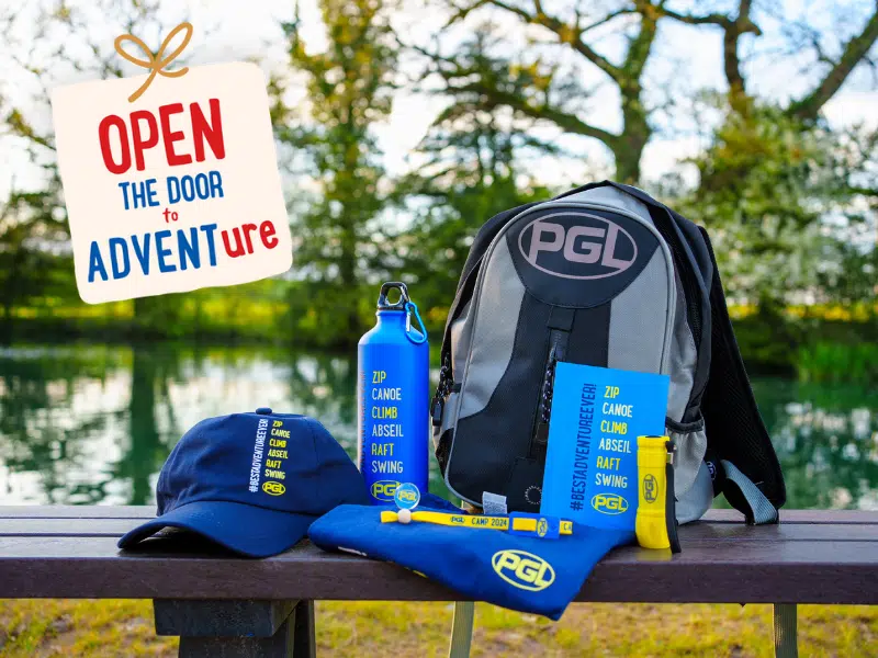 Outdoor adventure gear from PGL Adventure Holidays is displayed on a bench, including a backpack, cap, water bottle, and flyers. A sign by the lake reads 